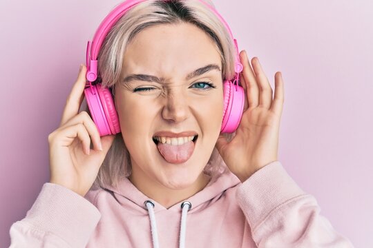 Young blonde girl listening to music using headphones sticking tongue out happy with funny expression.