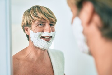 Handsome blond man looking at the mirror at the bathroom saving using foam and razor