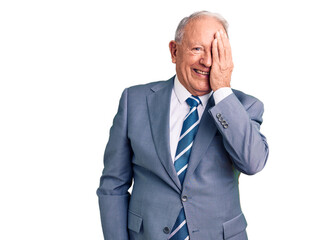 Senior handsome grey-haired man wearing elegant suit covering one eye with hand, confident smile on face and surprise emotion.