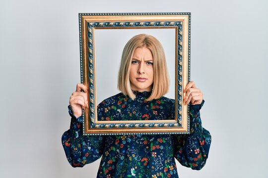 Young blonde woman holding empty frame clueless and confused expression. doubt concept.