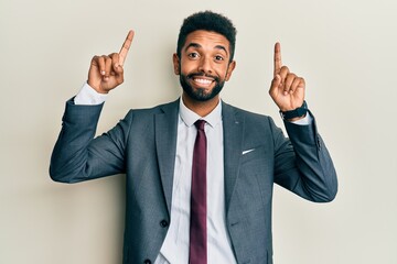 Handsome hispanic man with beard wearing business suit and tie smiling amazed and surprised and pointing up with fingers and raised arms.