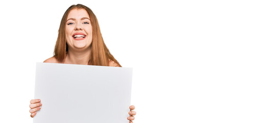 Young beautiful redhead woman holding blank empty banner looking positive and happy standing and smiling with a confident smile showing teeth