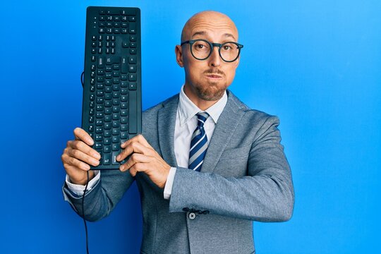 Bald man with beard holding keyboard puffing cheeks with funny face. mouth inflated with air, catching air.