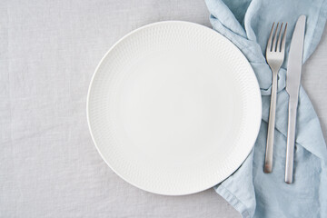 Clean empty white plate, fork and knife on pastel grey linen tablecloth on table, copy space, mock up, top view. Concept for menu with utensil
