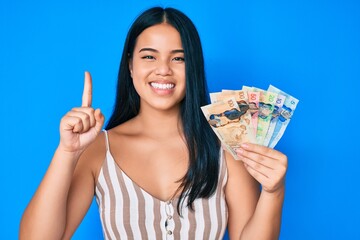 Young asian girl holding canadian dollars smiling with an idea or question pointing finger with happy face, number one