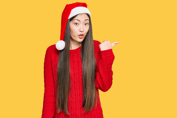 Young chinese woman wearing christmas hat surprised pointing with hand finger to the side, open mouth amazed expression.