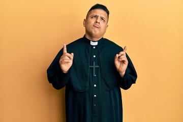 Young latin priest man standing over yellow background pointing up looking sad and upset,...