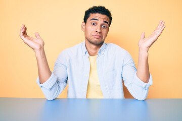 Young handsome hispanic man wearing casual clothes sitting on the table clueless and confused...