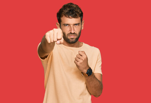 Handsome young man with beard wearing casual tshirt punching fist to fight, aggressive and angry attack, threat and violence
