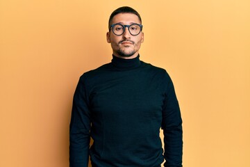 Handsome man with tattoos wearing turtleneck sweater and glasses relaxed with serious expression on face. simple and natural looking at the camera.