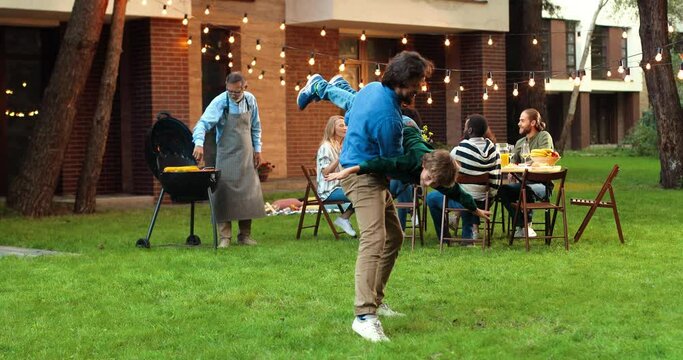 Caucasian cheerful father holding small cute son on hands, swinging and playing at back yard of house on weekend. Family at dinner table on background. Little boy play with dad and laughing laud.
