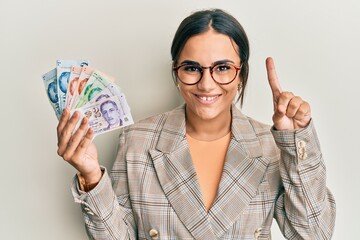 Young brunette woman holding singapore dollars banknotes smiling with an idea or question pointing finger with happy face, number one