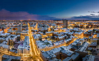 Naklejka premium Riga city panorama in winter time. Street lights glowing in blue hour. Snowy rooftops. Iconic buildings and architecture. Drone footage. 