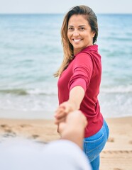 Beautiful middle age woman holding hands with husband at the beach, leading the way happy smiling in love