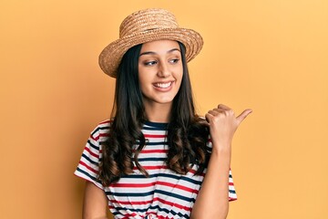 Young hispanic woman wearing summer hat smiling with happy face looking and pointing to the side with thumb up.