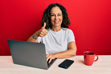 Beautiful middle age woman working at the office using computer laptop smiling happy and positive, thumb up doing excellent and approval sign