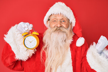 Old senior man with grey hair and long beard wearing santa claus costume holding alarm clock screaming proud, celebrating victory and success very excited with raised arm