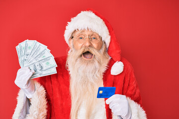Old senior man wearing santa claus costume holding dollars and credit card smiling and laughing hard out loud because funny crazy joke.