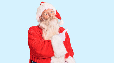 Old senior man with grey hair and long beard wearing traditional santa claus costume pointing with hand finger to the side showing advertisement, serious and calm face