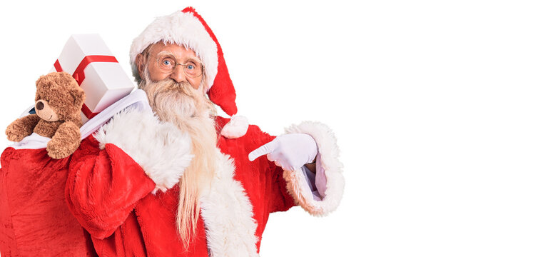 Old senior man with grey hair and long beard wearing santa claus costume holding bag with presents pointing finger to one self smiling happy and proud