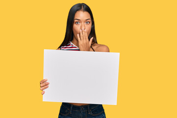 Beautiful hispanic woman holding blank empty banner covering mouth with hand, shocked and afraid for mistake. surprised expression