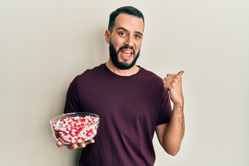 Young man with beard holding bowl full of pills pointing thumb up to the side smiling happy with open mouth