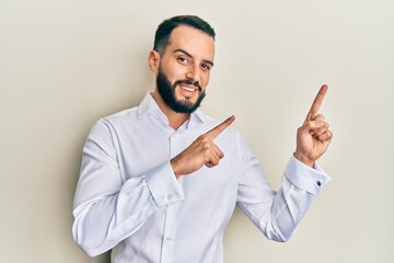 Young man with beard wearing business white shirt smiling and looking at the camera pointing with two hands and fingers to the side.