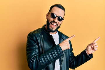 Young man with beard wearing black leather jacket and sunglasses smiling and looking at the camera pointing with two hands and fingers to the side.