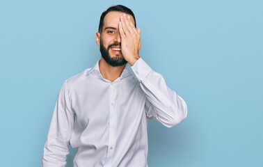 Young man with beard wearing business shirt covering one eye with hand, confident smile on face and surprise emotion.