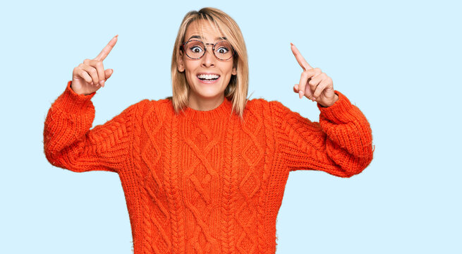 Beautiful blonde woman wearing casual clothes and glasses smiling amazed and surprised and pointing up with fingers and raised arms.