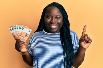 Young black woman with braids holding bunch of 50 euro banknotes smiling with an idea or question...