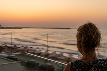 Fototapeta premium Blond girl with curly hair looking towards Old Venetian harbor and beach with umbrellas in the evening in city of Rethymno, Crete, Greece
