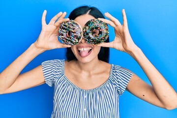 Beautiful young woman holding tasty colorful doughnuts on eyes sticking tongue out happy with funny expression.