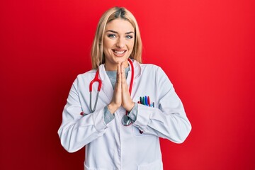 Young caucasian woman wearing doctor uniform and stethoscope praying with hands together asking for...