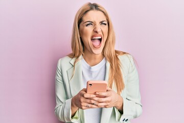 Young caucasian woman wearing business clothes using smartphone angry and mad screaming frustrated and furious, shouting with anger. rage and aggressive concept.