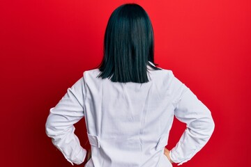 Young hispanic woman wearing doctor uniform and stethoscope standing backwards looking away with arms on body