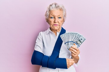 Senior grey-haired woman wearing arm on sling holding usa dollars banknotes relaxed with serious expression on face. simple and natural looking at the camera.