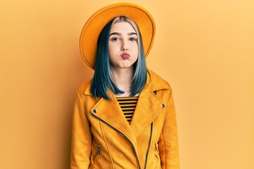 Young modern girl wearing yellow hat and leather jacket puffing cheeks with funny face. mouth inflated with air, crazy expression.