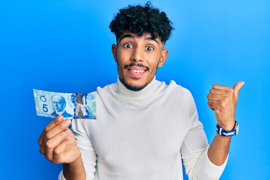 Young arab handsome man holding 5 canadian dollars banknote pointing thumb up to the side smiling happy with open mouth
