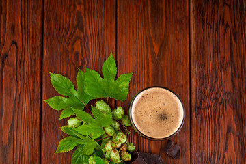 Fototapeta na wymiar Glass of dark stout beer with natural green hop leaves and cones and chopped chocolate on a wooden table. Top view. Text space.