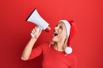 Young blonde woman wearing christmas hat screaming using megaphone over isolated red background.