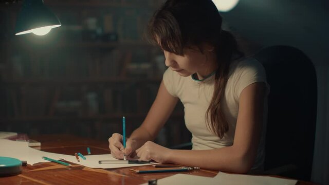 Inspired child draws with enthusiasm in evening by light of table lamp. Little girl schoolgirl draws with pencils doing homework