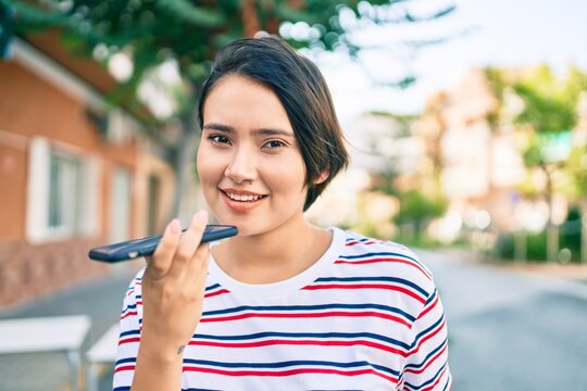 Young latin girl smiling happy sending voice message using smartphone at the city