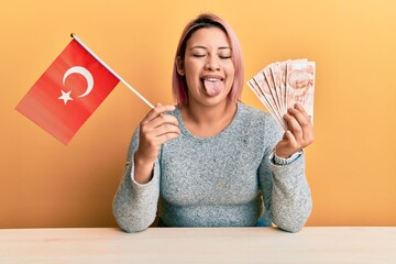 Hispanic woman with pink hair holding turkey flag and liras banknotes sticking tongue out happy with funny expression.