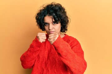 Fototapeta na wymiar Young hispanic woman with curly hair wearing casual winter sweater ready to fight with fist defense gesture, angry and upset face, afraid of problem