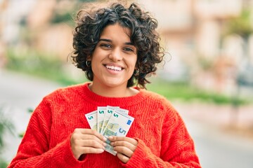 Young hispanic woman smiling happy holding 5 euro banknotes at the city.