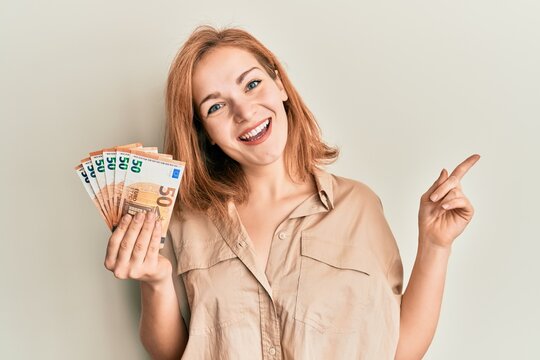 Young caucasian woman holding bunch of 50 euro banknotes smiling happy pointing with hand and finger to the side