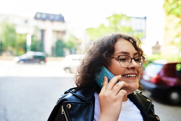 Smiling curly woman wearing trendy sunglasses walks down the central city street and uses her phone.