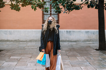 Young blonde woman with face mask and colorful shopping on an orange wall in the street.