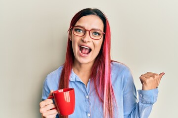 Young caucasian woman holding coffee pointing thumb up to the side smiling happy with open mouth